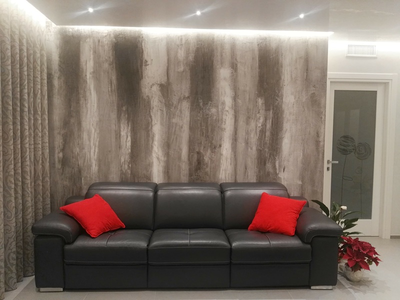 Soggiorno con parete in finitura decorativa grigia sfumata, divano in pelle nera con cuscini rossi e luce LED nel controsoffitto